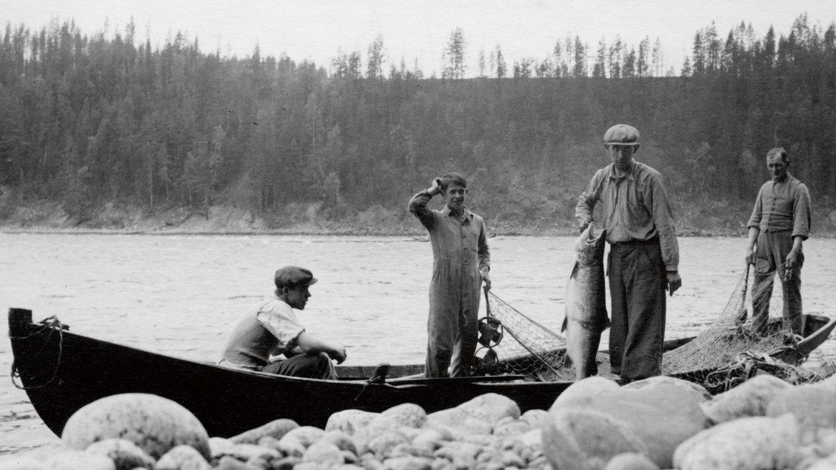 En svartvit bild på fyra personer vid en båt. De har nät i båten, och en av dem håller i en stor lax.